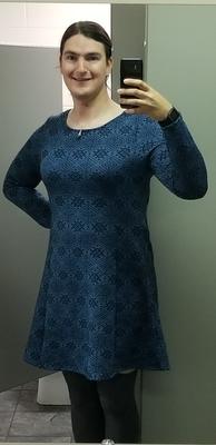 A woman takes a selfie in an office bathroom. She is smiling, and has her dark hair pulled into a low ponytail with a bow barrette at her right temple. She is wearing a long-sleeved teal dress with a geometric pattern in black printed on it. She is also wearing comfy thigh-high grey socks.