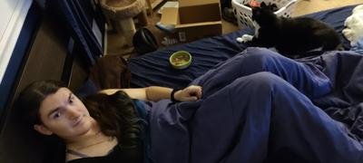 A solemn woman takes a selfie in her bed. She is wearing her long dark hair in a ponytail over her right shoulder, showing that the lower third is dyed deep blue. She has a black camisole one, and a tuxedo cat named Moxie on her bed with a bowl of food.